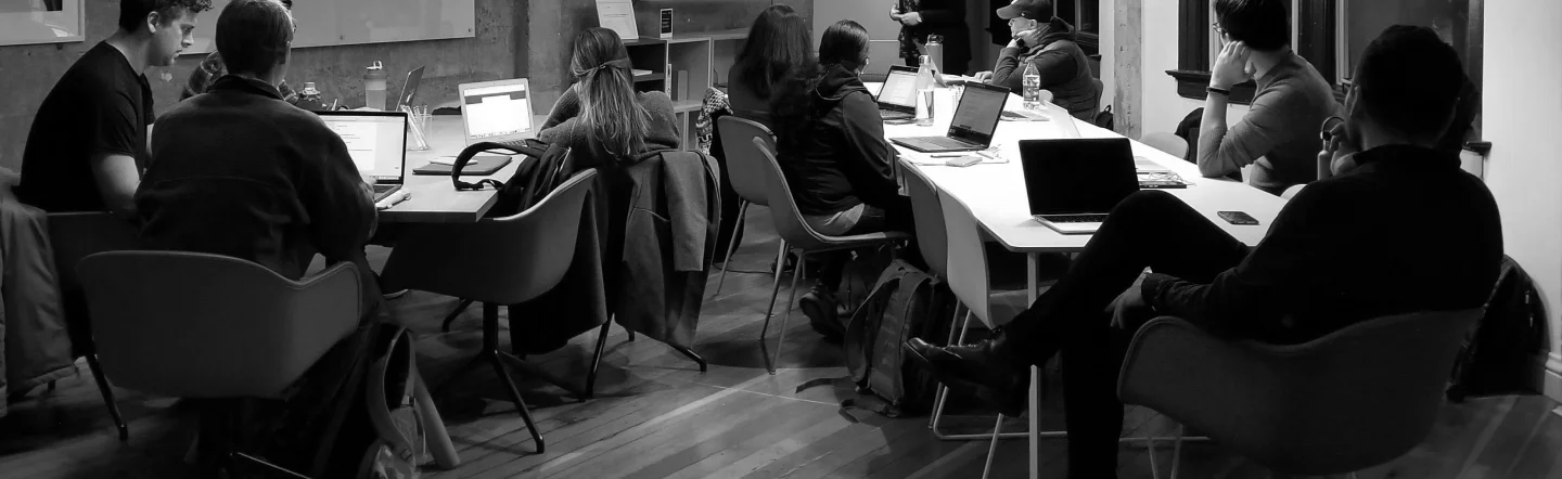 Black and white photo of a group of people seated around a long table working on laptops in a modern office setting.